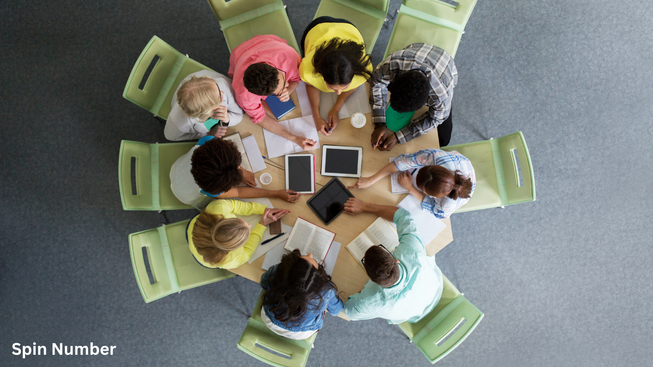 A top-down bird's-eye view of eight people sitting in a circle at a round wooden table working with tablets and books, highlighting a group sorted by a random team generator.