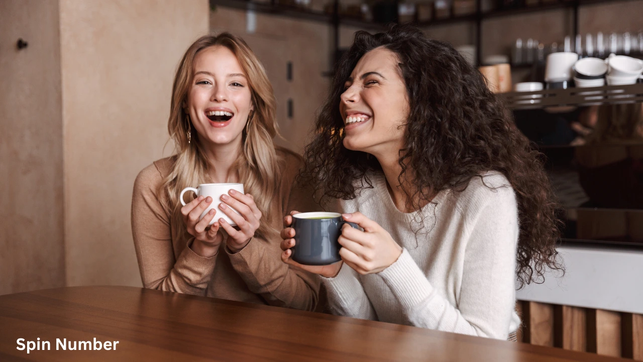 Two young women sitting at a wooden cafe table laughing hysterically over coffee mugs, showcasing a great environment for sharing fun Questions to Ask Your Crush.