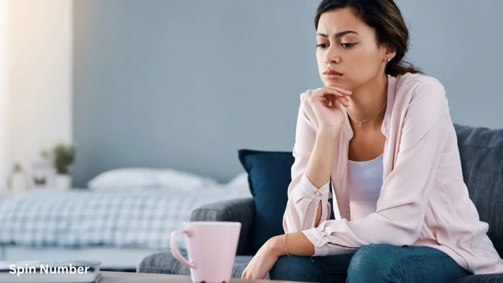 A worried young woman sitting on a living room couch with her hand on her chin, staring blankly ahead as she struggles with How to Stop Overthinking.