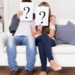 A young man and woman sitting together on a white couch holding up paper sheets with large question marks over their faces, symbolizing the search for deep Questions for Couples.