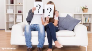 A young man and woman sitting together on a white couch holding up paper sheets with large question marks over their faces, symbolizing the search for deep Questions for Couples.