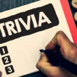 A close-up of a person writing in a spiral notebook labeled "TRIVIA" next to a numbered list, preparing a set of Trivia Questions.
